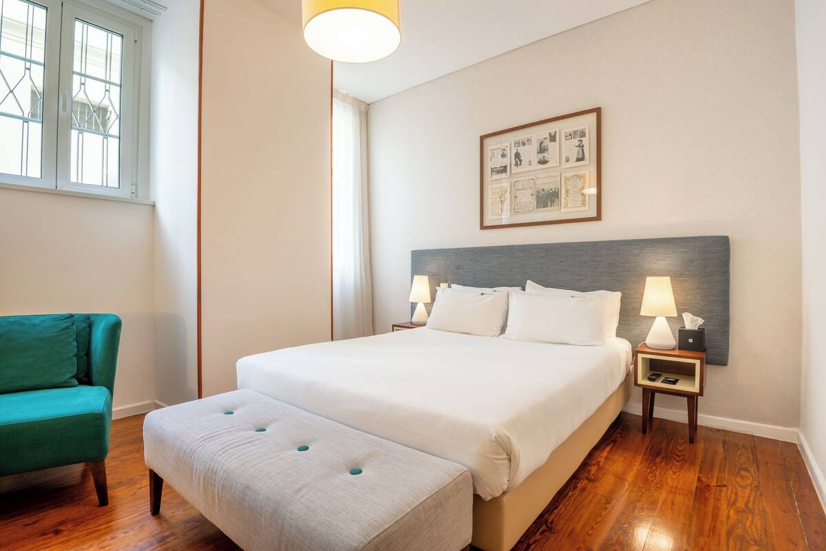 Habitación de hotel ordenada con una cama grande, un sillón verde, una mesita de noche con una lámpara y un cuadro en la pared.