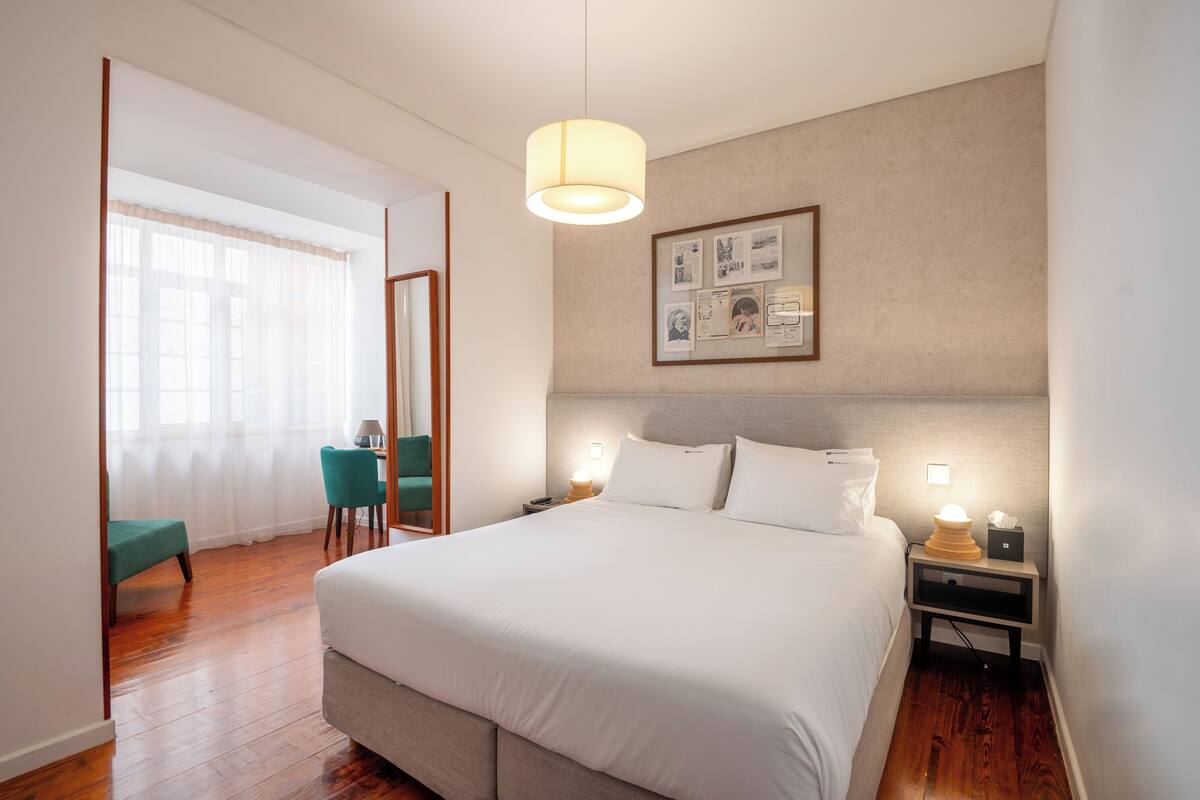 Una habitación de hotel con una cama grande, mesitas de noche, un escritorio con silla y una ventana con cortinas translúcidas.