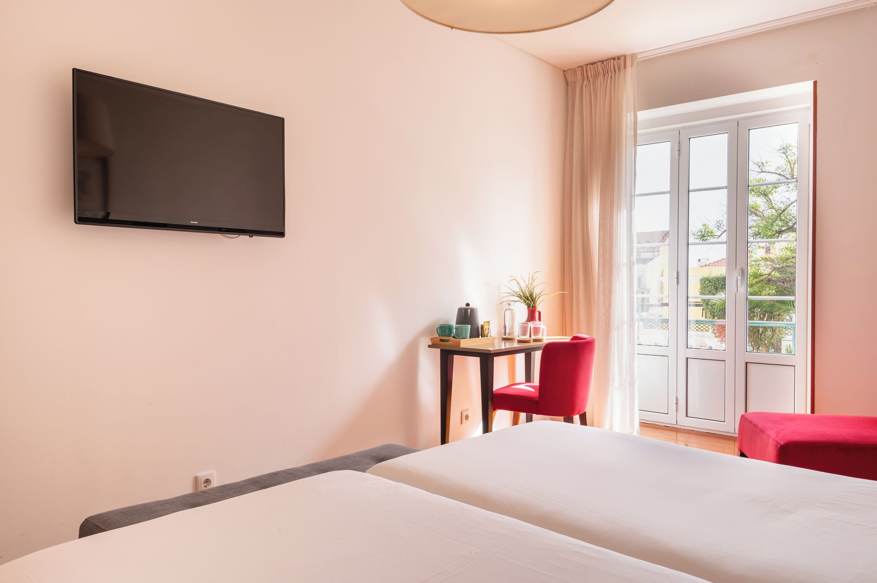 Een hotelkamer met een bed, een aan de muur bevestigde televisie, een klein tafeltje met een stoel, en uitzicht op de omgeving door een schuifglazen deur.