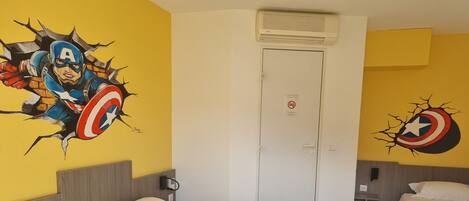 Habitación de hotel con una cama grande, dos camas más pequeñas, una puerta con letrero de no fumar y una pared con un mural de Captain America.
