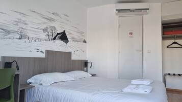 A hotel room with a bed, a green chair, a wall mural, an air conditioning unit, and a door with a no-smoking sign.