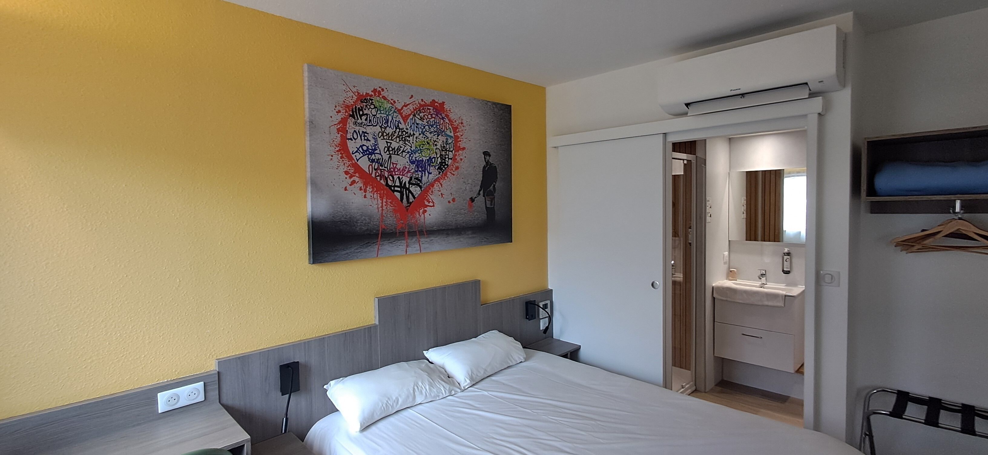 A hotel room with a bed, a bedside table, a wall-mounted TV, a bathroom with a mirror, and a door leading to another room.