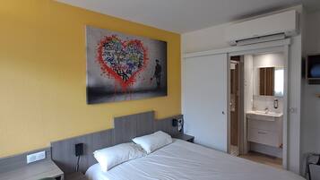 A hotel room with a bed, a bedside table, a wall-mounted TV, a bathroom with a mirror, and a door leading to another room.