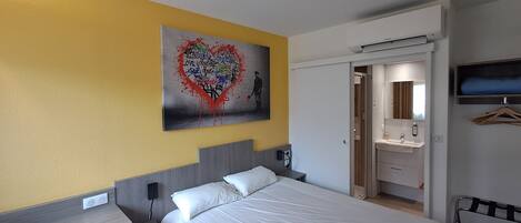 A hotel room with a bed, a bedside table, a wall-mounted TV, a bathroom with a mirror, and a door leading to another room.