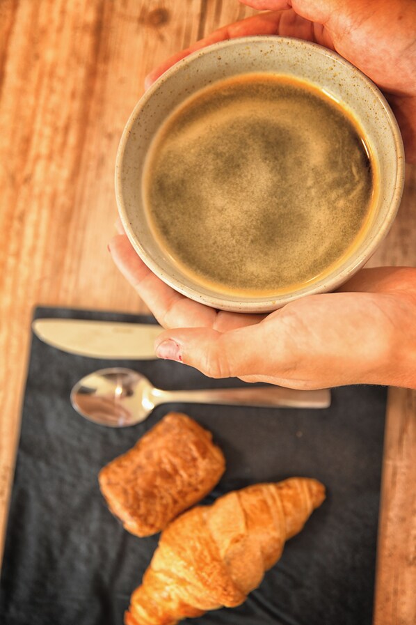 Una persona sostiene una taza de café, una cuchara y dos cruasanes sobre una superficie oscura.