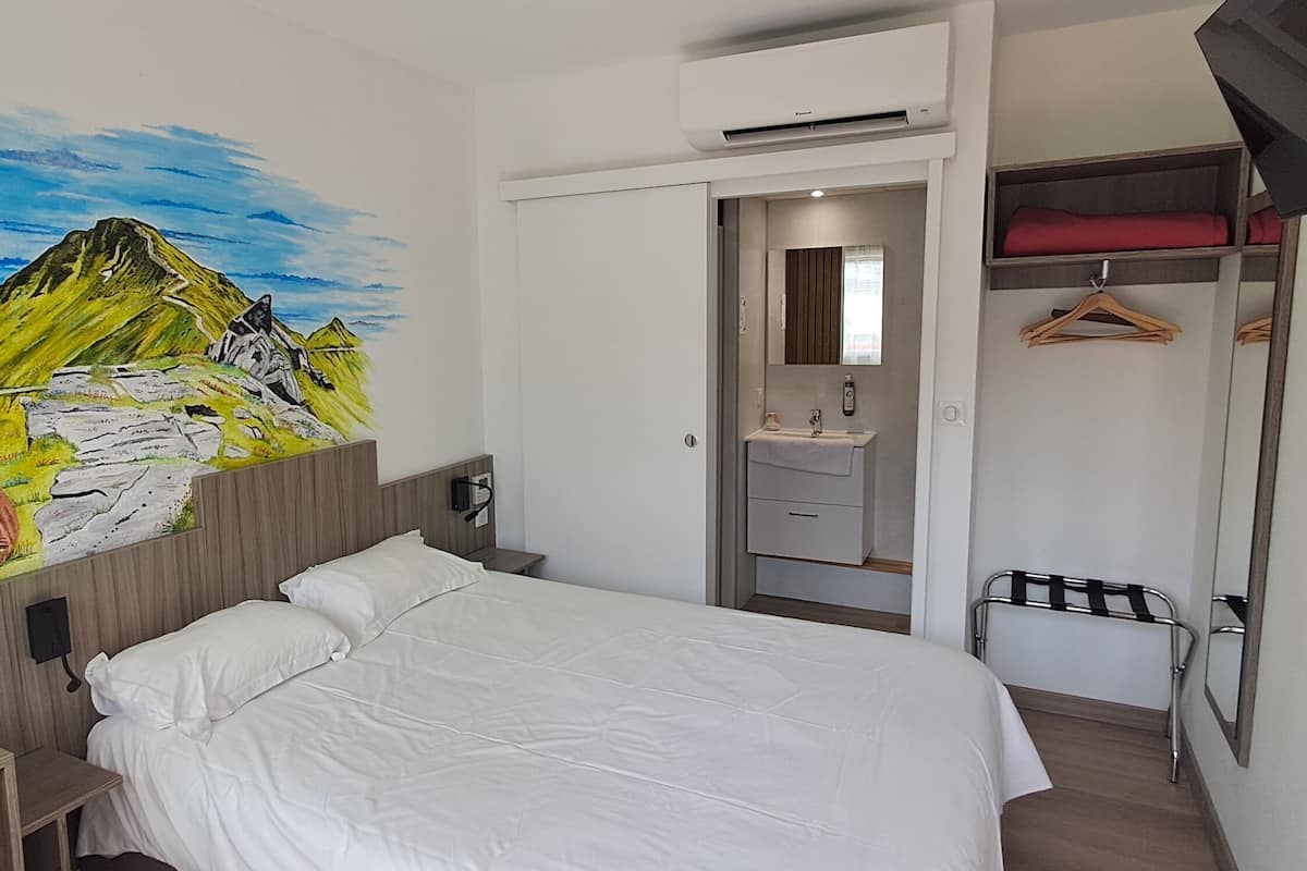 Ein Schlafzimmer mit Bett, Schreibtisch, Schminktisch, Spiegel, Kleiderhaken und Wandbild einer Berglandschaft.