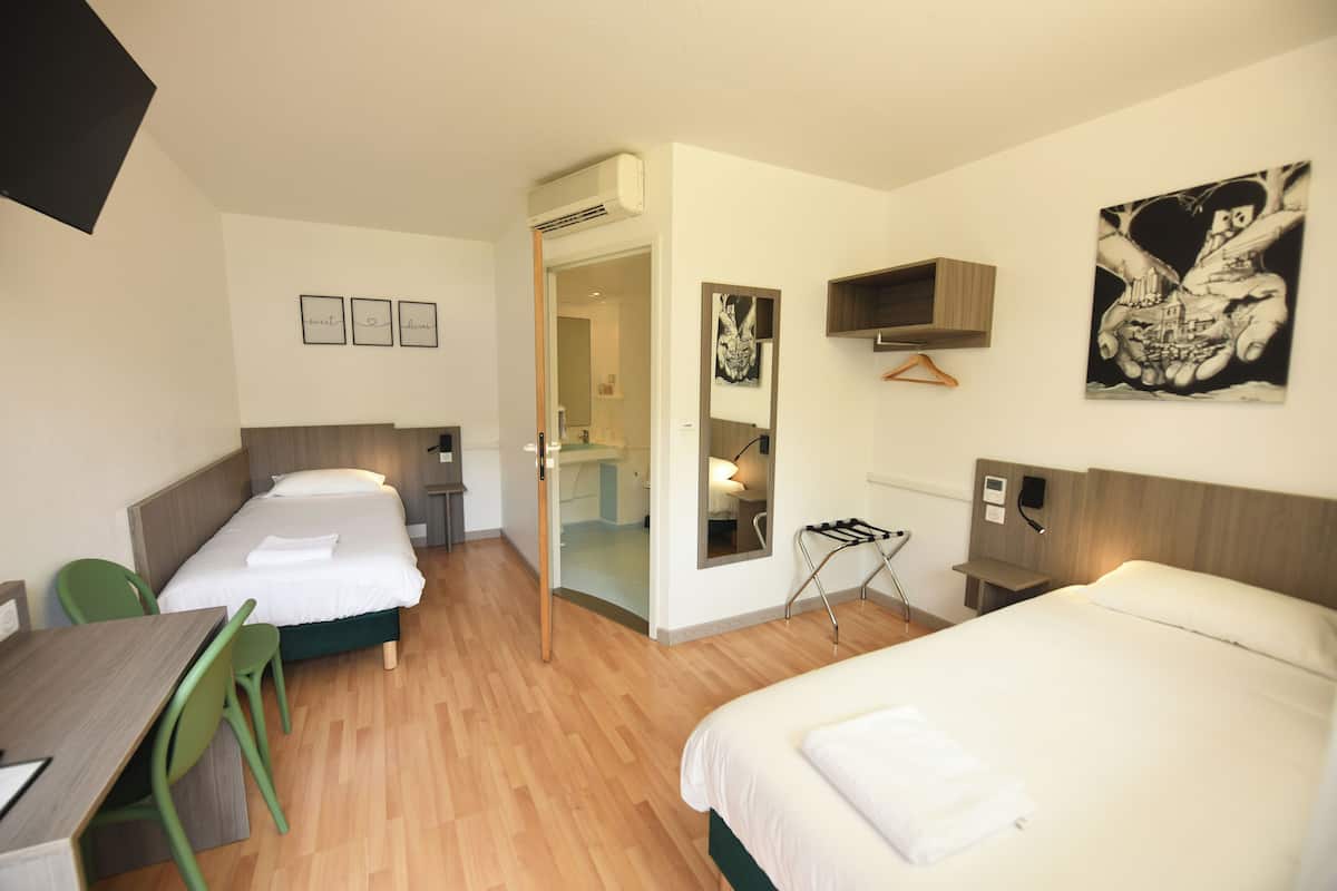 Ein Hotelzimmer mit zwei Betten, einem Schreibtisch und einem Sessel. Man sieht einen Spiegel, einen Bilderrahmen und eine Pendelleuchte.