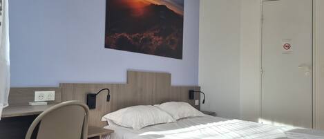A hotel room with a bed, a desk, a chair, a wall-mounted painting, and a door with a no-smoking sign.