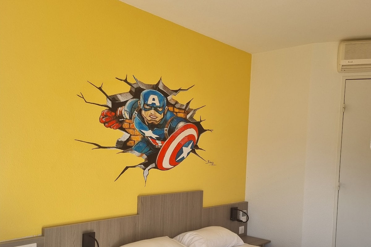 Ein Hotelzimmer mit Bett, Schreibtisch und Wandbild von Captain America.