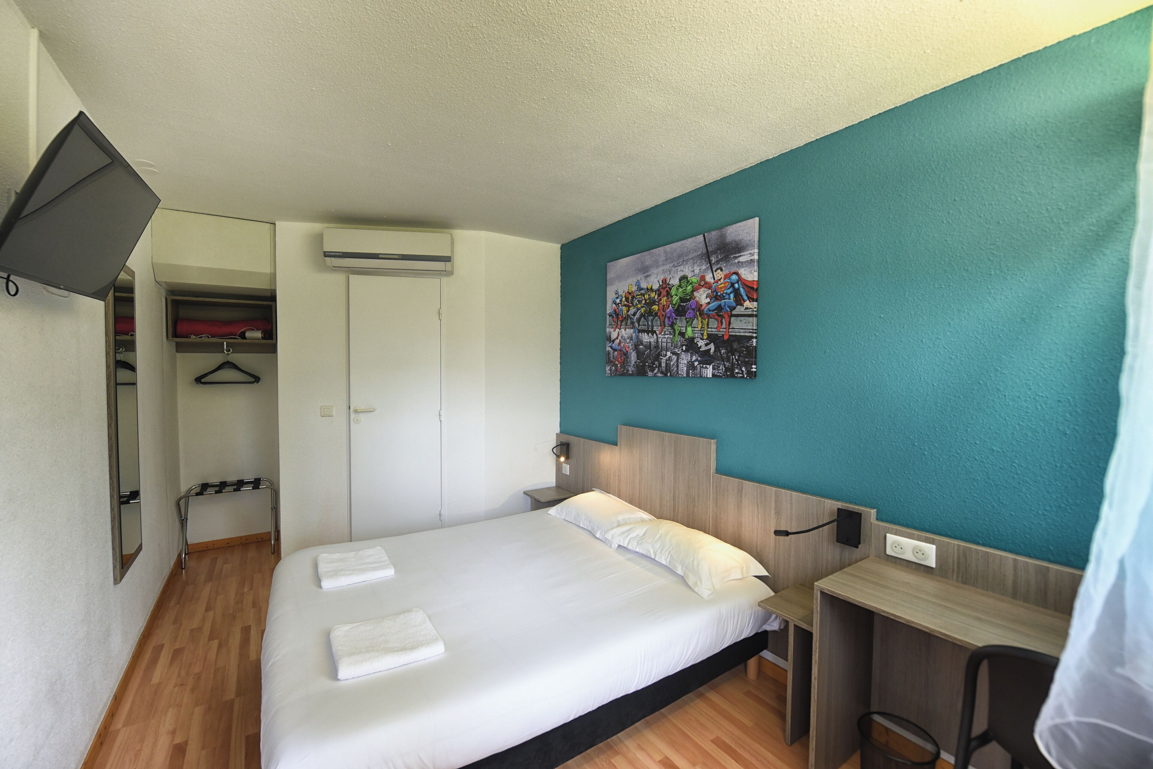 A hotel room with a bed, a desk, a chair, a wardrobe, and a painting on the wall.