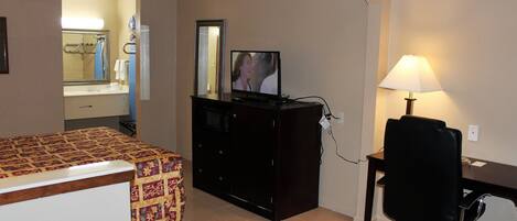 A hotel room with a bed, a desk with a computer, a TV, a lamp, and a bathroom visible through an open door.