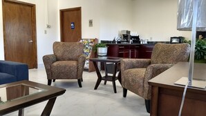 Un salon avec deux fauteuils à motifs, une table basse en verre et un canapé bleu. On aperçoit à l’arrière-plan un coin cuisine avec une machine à café et des placards.