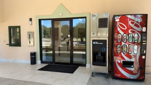 Une entrée d’immeuble avec une porte vitrée, un distributeur de glaçons à l’effigie de Coca-Cola et une poubelle.
