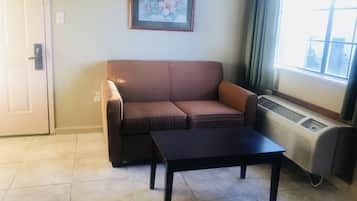 A room with a brown sofa, a black table, a framed floral picture on the wall, and a window with curtains.
