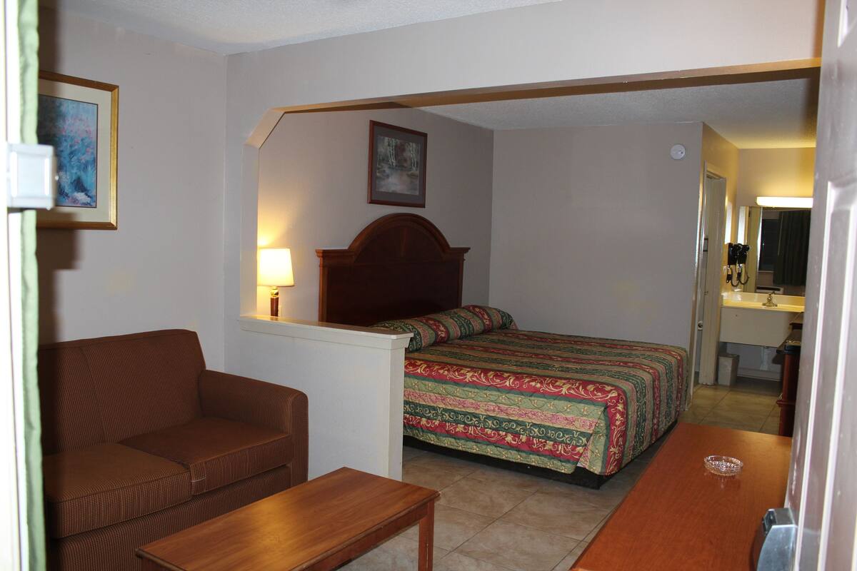 A hotel room with a bed, a sofa, a wooden table, and a small bathroom visible through an open door.