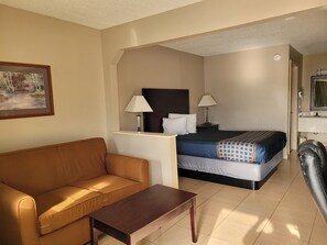 A hotel room with a bed, a sofa, a small table, two bedside lamps, a painting on the wall, and a bathroom visible through an open door.