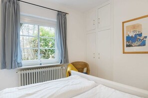 Une chambre à coucher avec un lit, une fenêtre avec des rideaux, un radiateur, une chaise et un tableau encadré accroché au mur.