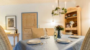 A dining area with a table set for four, surrounded by wicker chairs, a wooden cabinet with decorative items, and a chandelier. - House 7 "Beach Pirate" (ID 023) (Room category) - House 7 "Beach Pirate" (ID 023) (Sankt Peter-Ording)