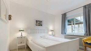 A bedroom with a white bed, two bedside tables with lamps, a radiator, a window with curtains, and a framed picture on the wall. - House 7 "Beach Pirate" (ID 023) (Room category) - House 7 "Beach Pirate" (ID 023) (Sankt Peter-Ording)