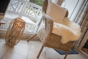 A wicker chair with a fluffy white cushion, a woven floor lamp, and a view of a white fence and greenery outside. - House 7 "Beach Pirate" (ID 023) (Room category) - House 7 "Beach Pirate" (ID 023) (Sankt Peter-Ording)