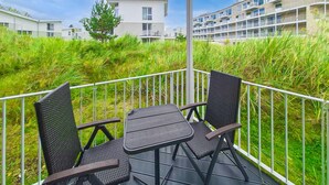 A balcony with two chairs and a table, surrounded by a white railing, overlooking a grassy area and modern buildings. - Dünenvilla 18, Whg 3 (Travemünde Waterfront)
