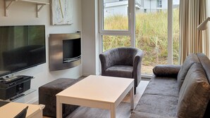 A modern living room with a flat-screen TV, a comfortable sofa, a grey armchair, and a small white table. - Dünenvilla 18, Whg 3 (Travemünde Waterfront)
