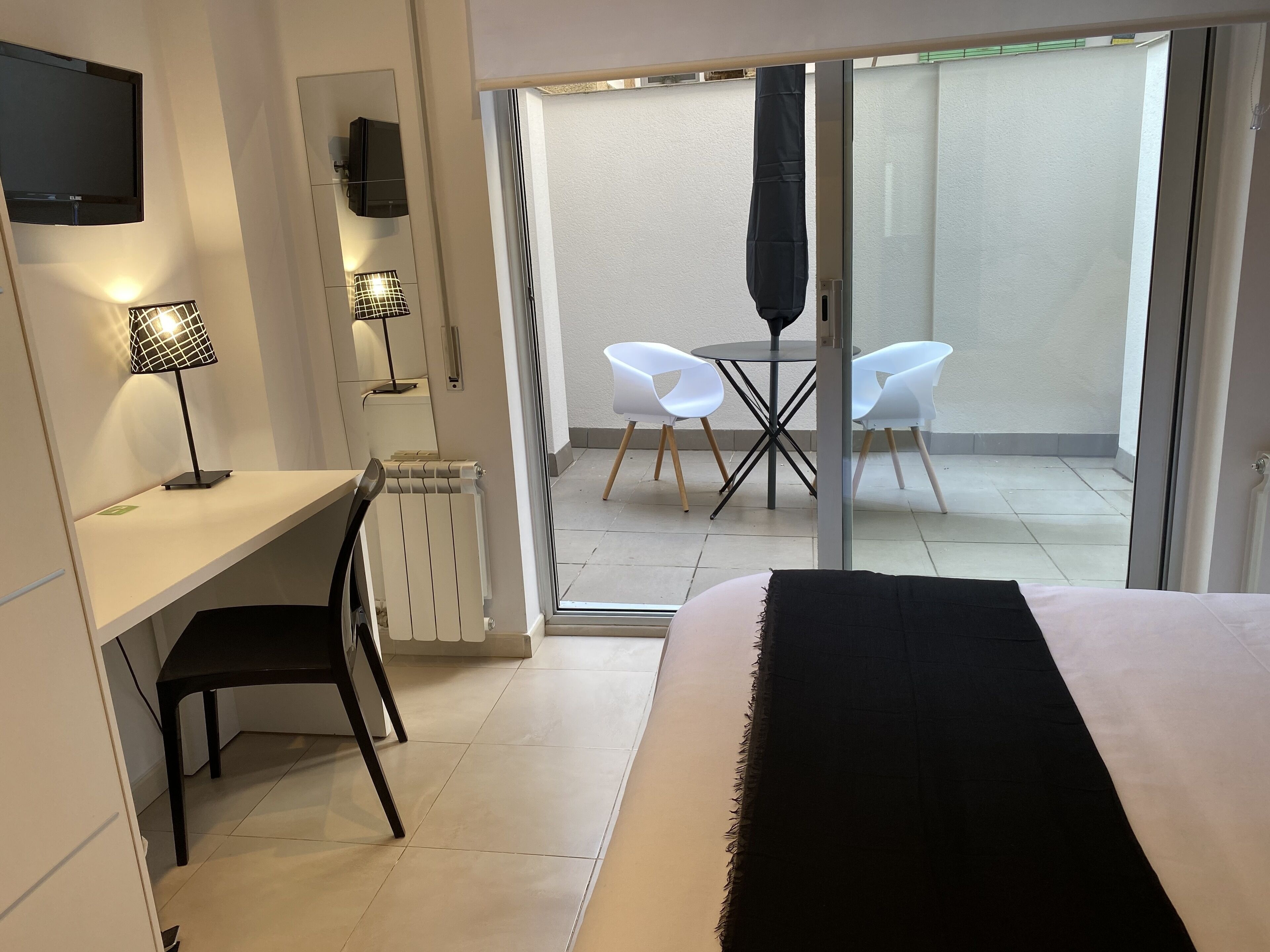 Habitació d'hotel amb llit, escriptori amb cadira, televisió, làmpada i vista a un balcó amb cadires i taula.