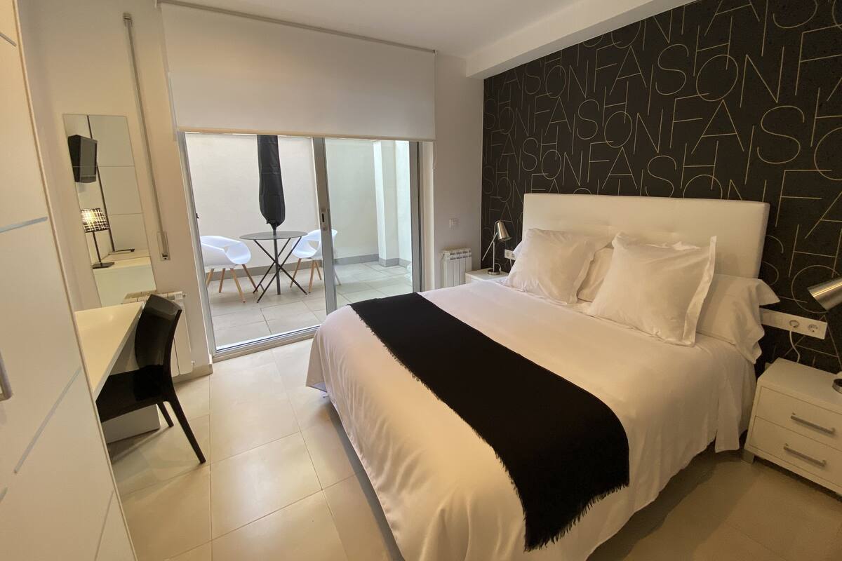 Una habitación de hotel moderna con una cama grande, un escritorio y una silla. Cuenta con un balcón con mesa y sillas.