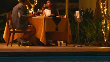 Una pareja cenando en una mesa al aire libre, con luces colgantes, una piscina y plantas de fondo.