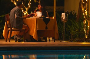 Una pareja cenando en una mesa al aire libre, con luces colgantes, una piscina y plantas de fondo.