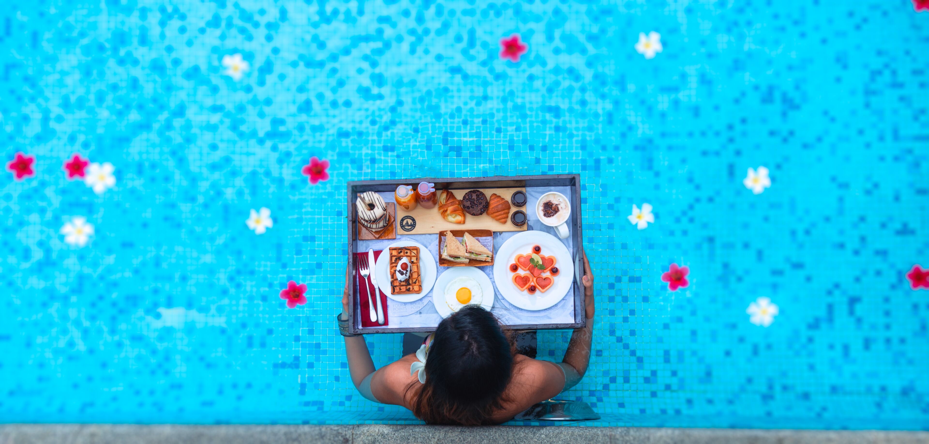 A person holding a tray with various food items by a pool with a blue tiled background.