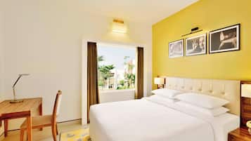 Habitación de hotel con cama, escritorio, silla, ventana con cortinas e imágenes enmarcadas en la pared.