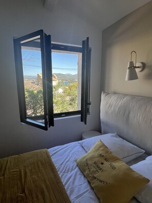 A bedroom with a bed, a window with open shutters, a wall-mounted lamp, and a pillow with text.