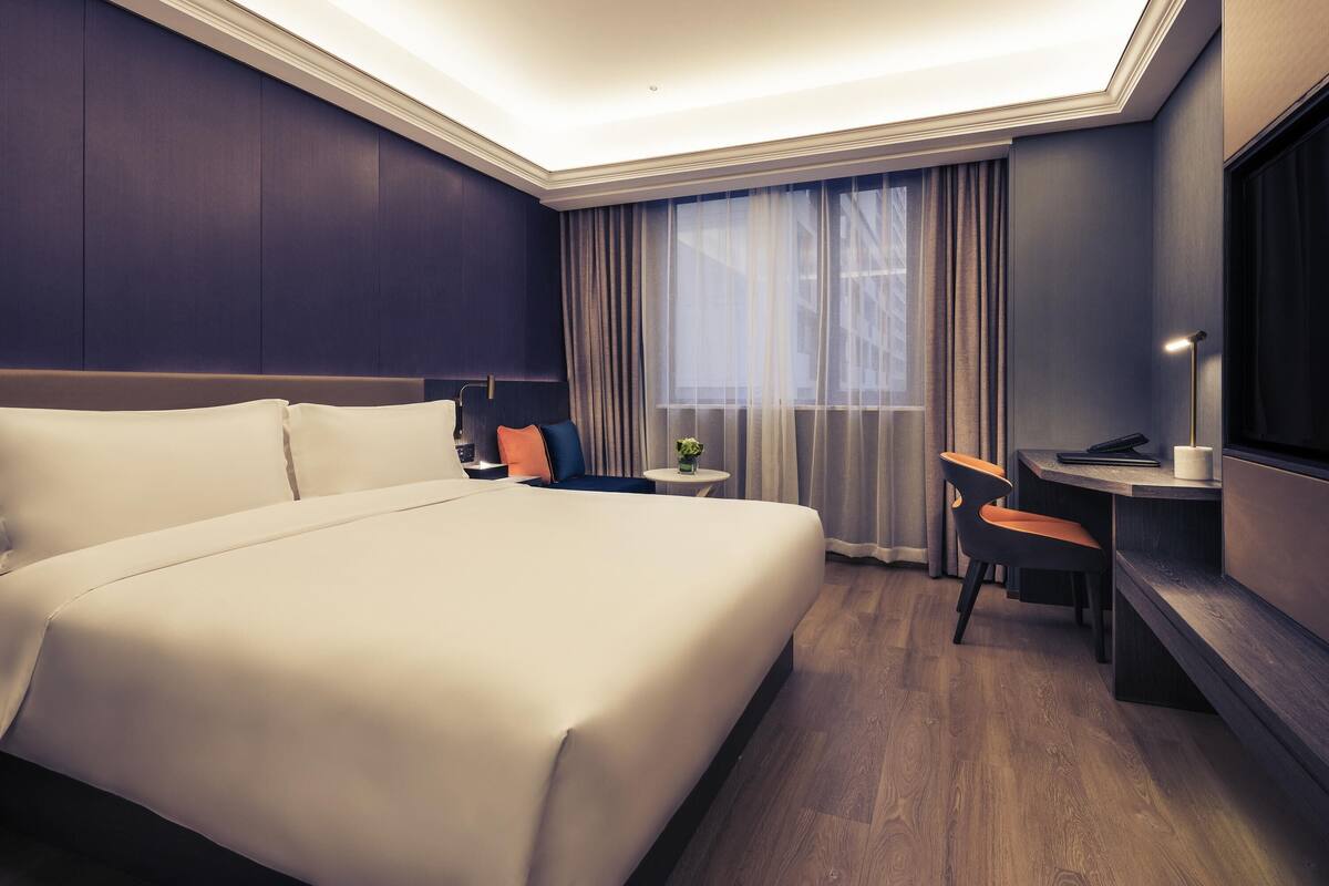 A hotel room with a large bed, a desk with a chair, a small table with a lamp, and a large window with curtains.