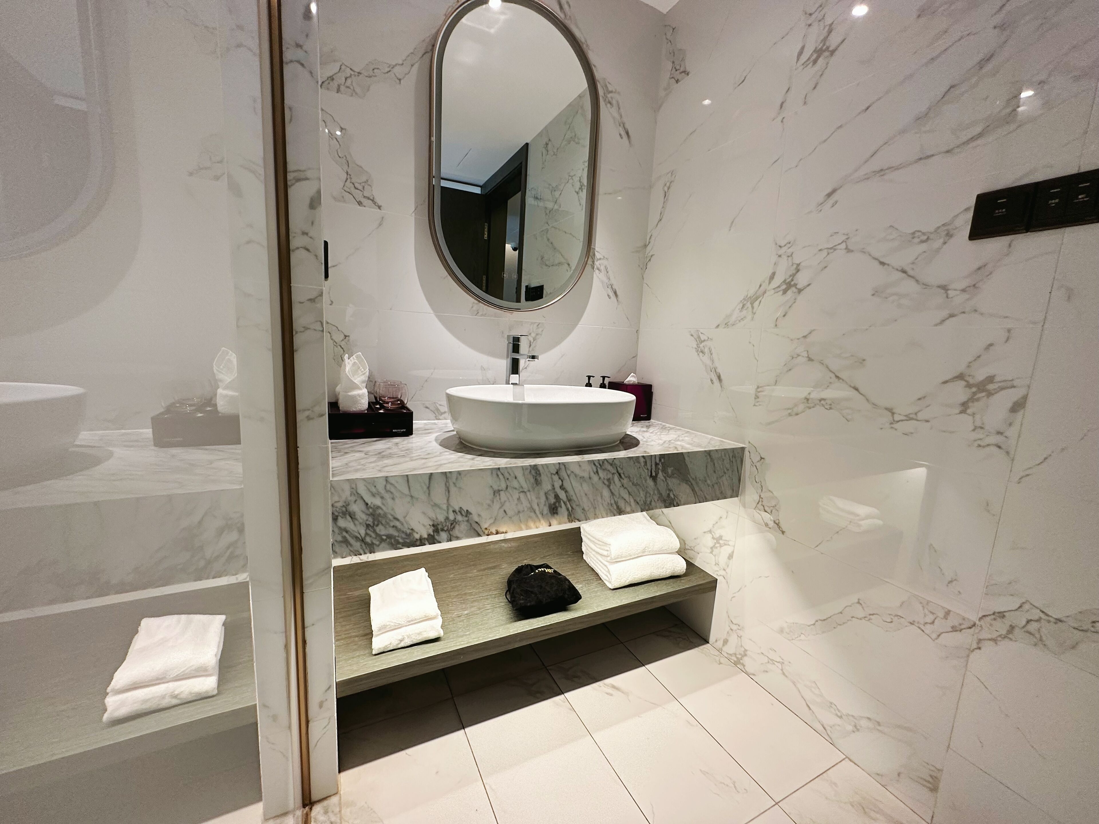 Une salle de bain moderne avec un grand miroir ovale, un lavabo rond, des comptoirs en marbre et des serviettes soigneusement pliées.