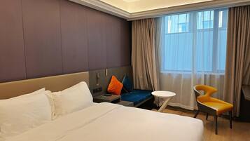 A hotel room with a large bed, a small table, a chair, and a window with curtains.