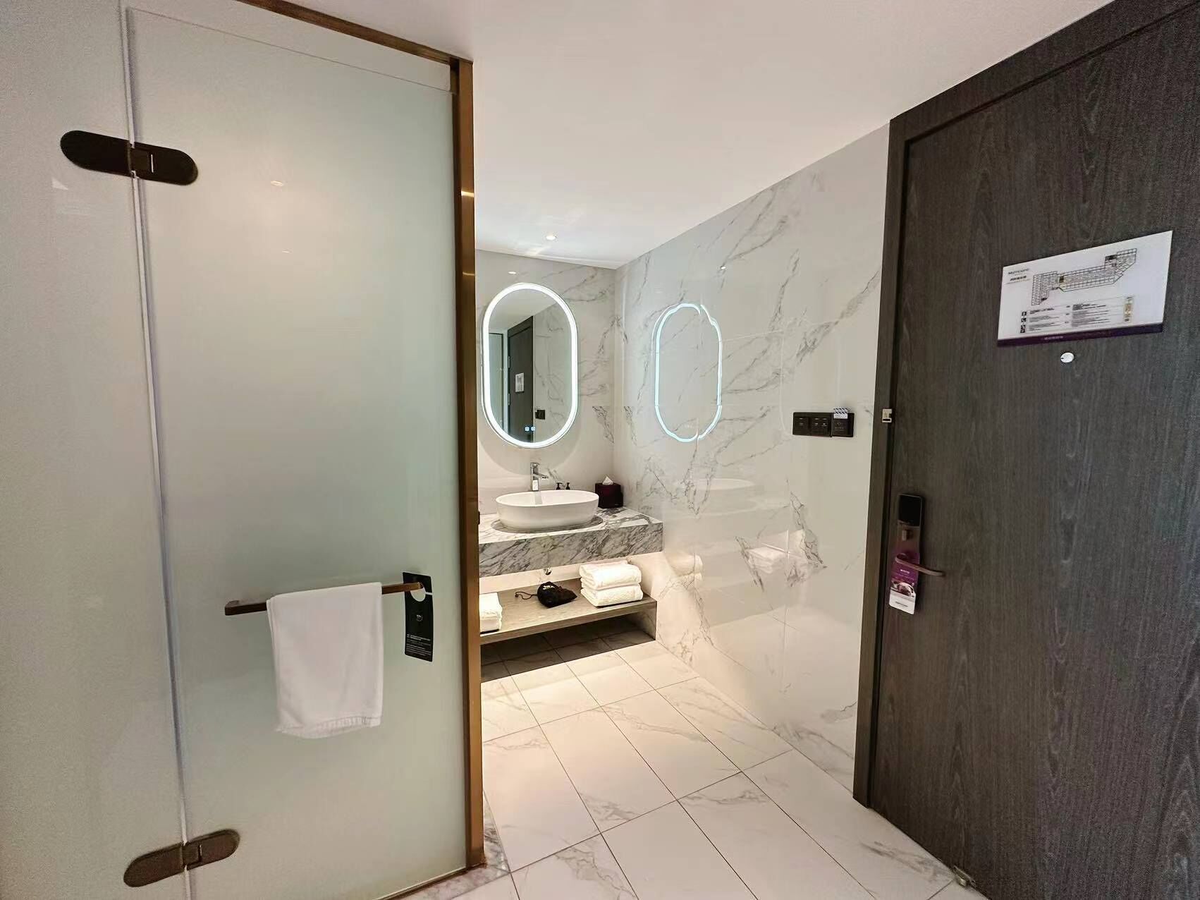 A modern bathroom with a glass door, a marble sink, and a large mirror with illuminated edges.