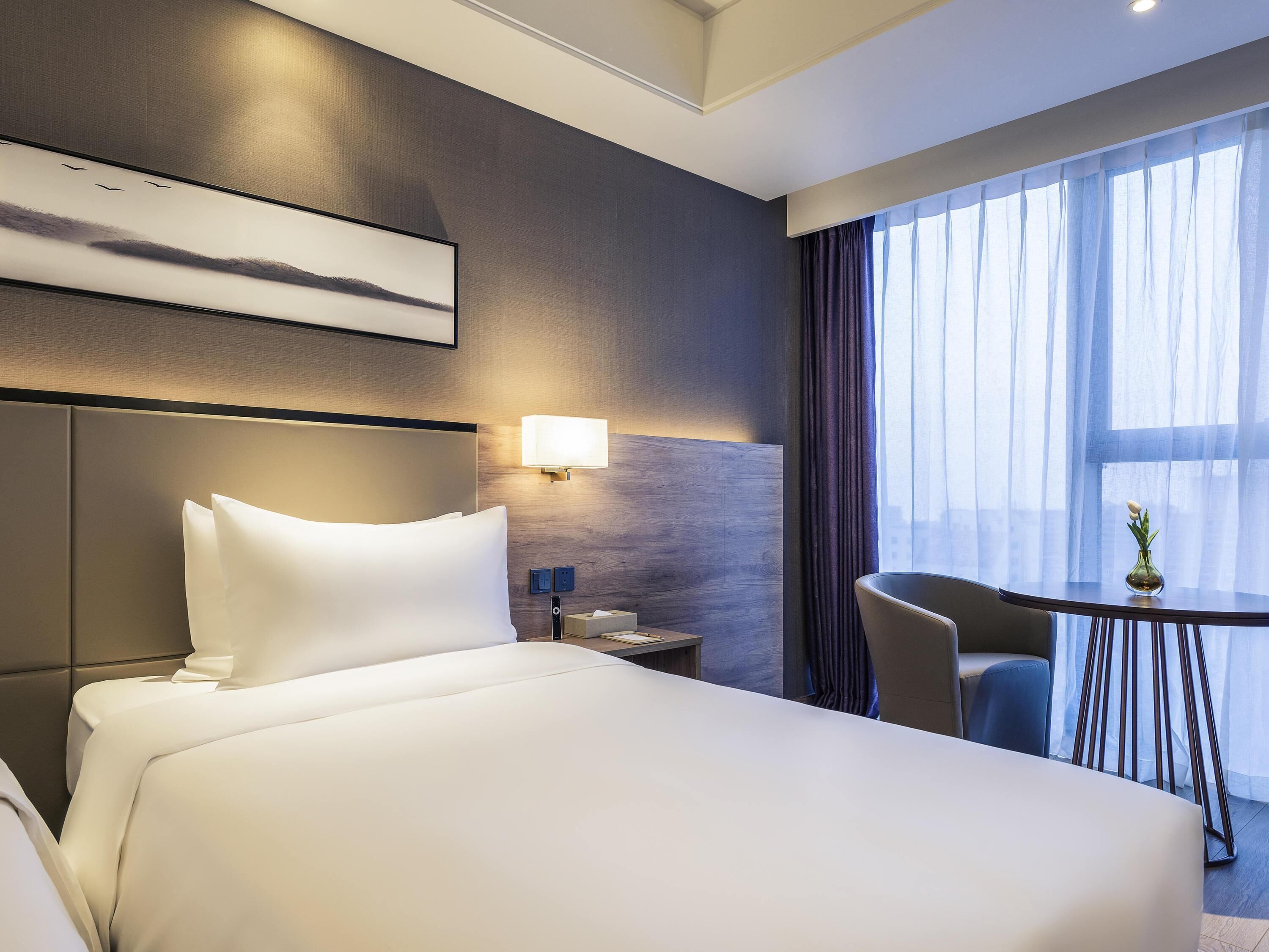 A hotel room with a large bed, a bedside table, a lamp, a chair, and a small table with a vase.