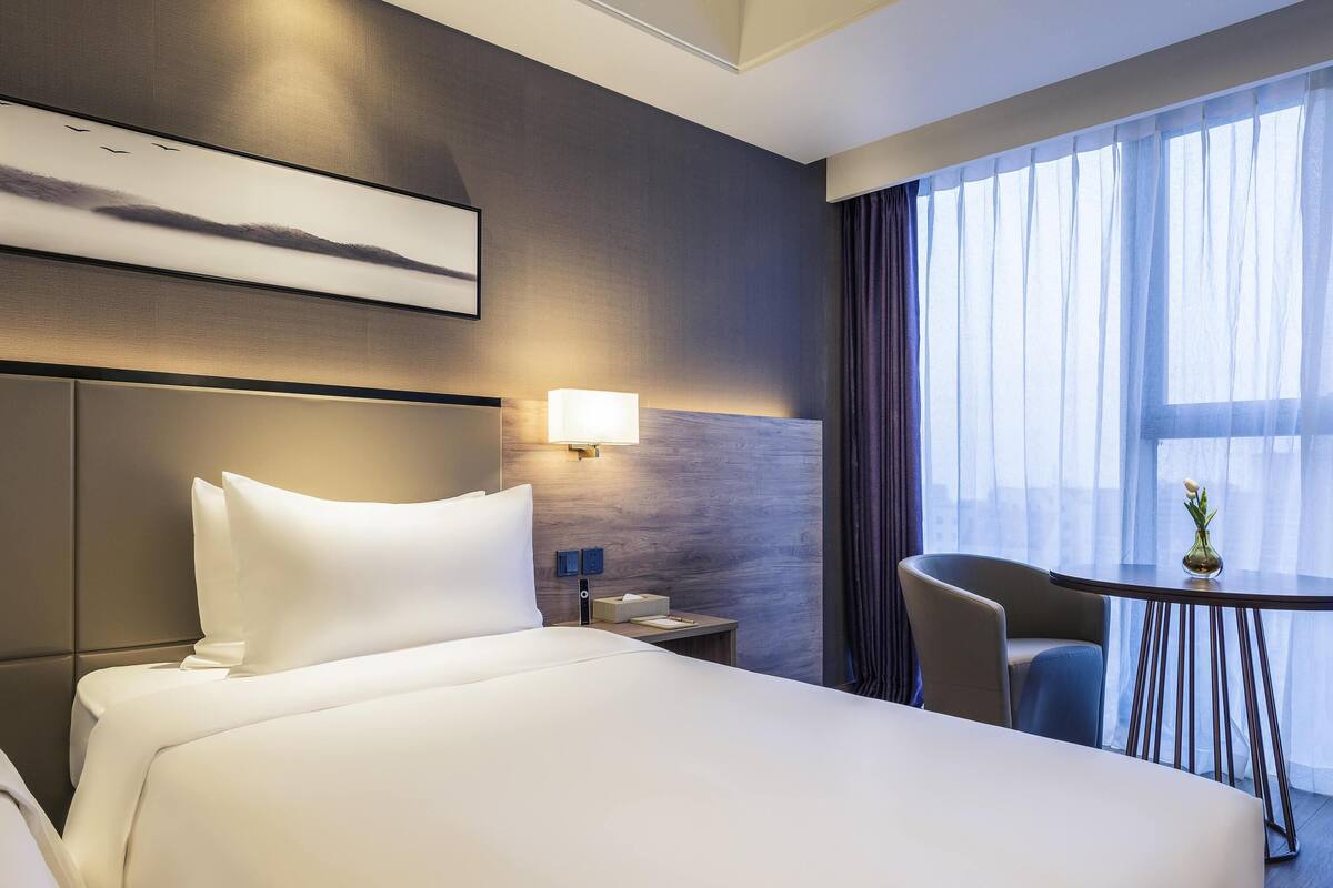 Hotellihuone, jossa on suuri sÀnky, yöpöytÀ, lamppu, tuoli ja pieni pöytÀ, jonka pÀÀllÀ on maljakko.