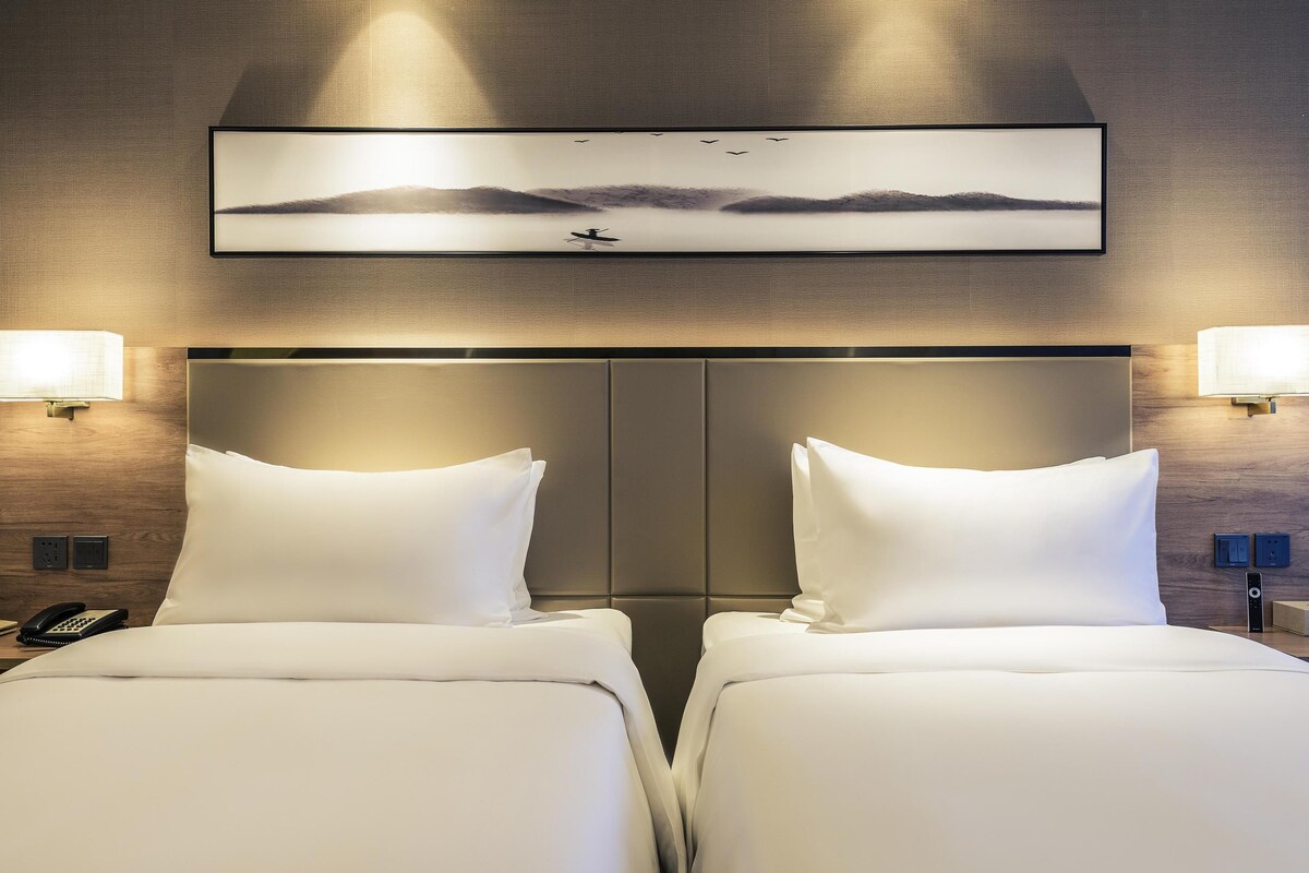 A hotel room with two beds, a large headboard, wall-mounted lamps, and a framed picture above the beds.