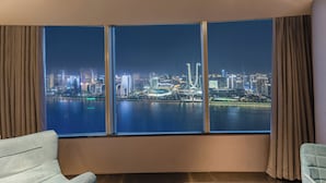 Een kamer met een groot raam, uitzicht op een verlicht stadsbeeld 's nachts, een houten vloer en twee lichte stoelen.