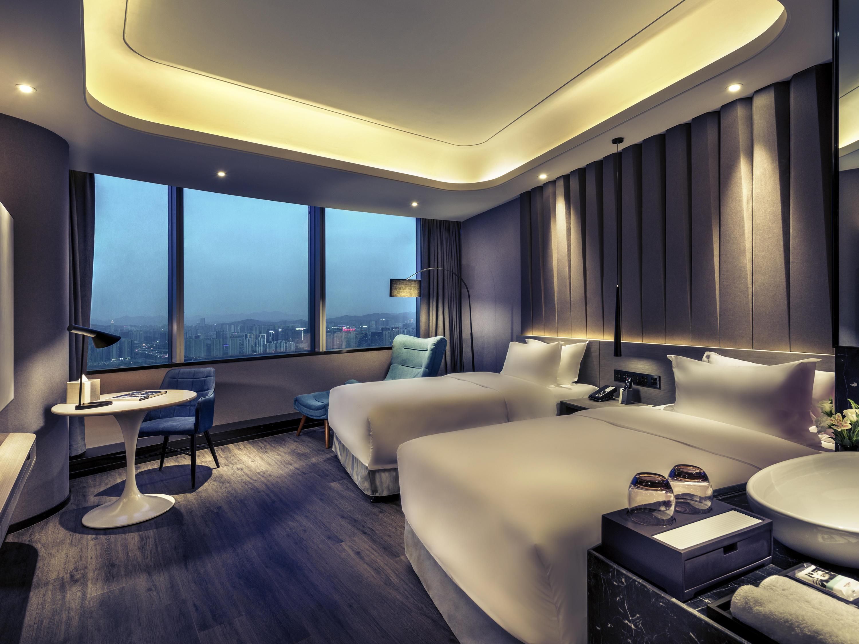 A modern hotel room with a large bed, a seating area with a chair and table, a bedside table with a lamp, and a view of the cityscape through large windows.