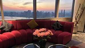 A red sofa with patterned pillows, a vase with pink flowers, and a view of a city skyline through large windows. - Mercure Hangzhou Qianjiang (Hangzhou)