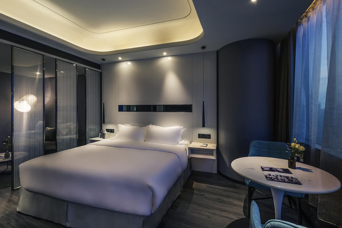 Habitació d'hotel moderna amb un llit gran, taules de nit, una taula rodona petita amb un ram i vista a la ciutat a través de cortines translúcides.