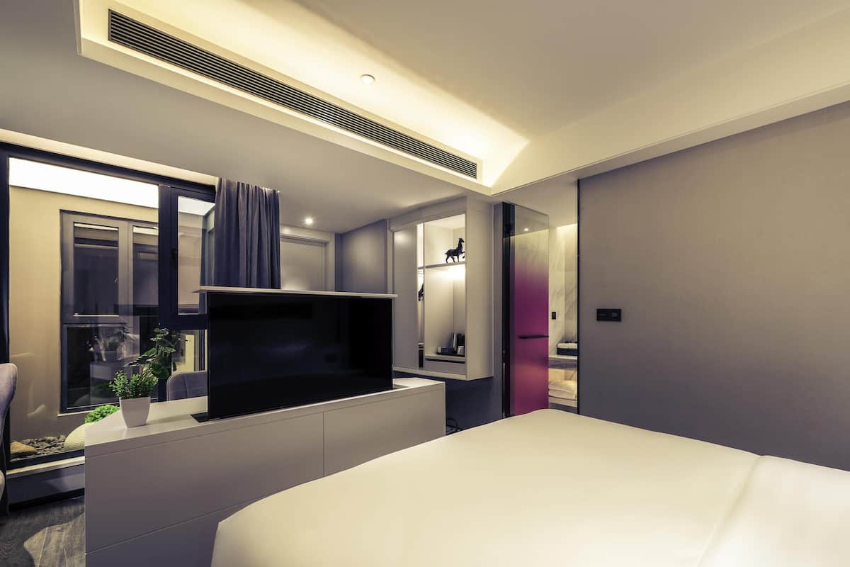 Une chambre d’hôtel moderne équipée d’un lit, d’un téléviseur à écran plat, d’un bureau et d’une salle de bain visible à travers une paroi en verre.