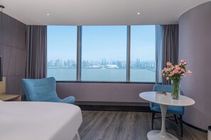 A hotel room with a large window, a bed, a blue armchair, a small round table, and a vase with flowers. - Mercure Hangzhou Qianjiang (Hangzhou)