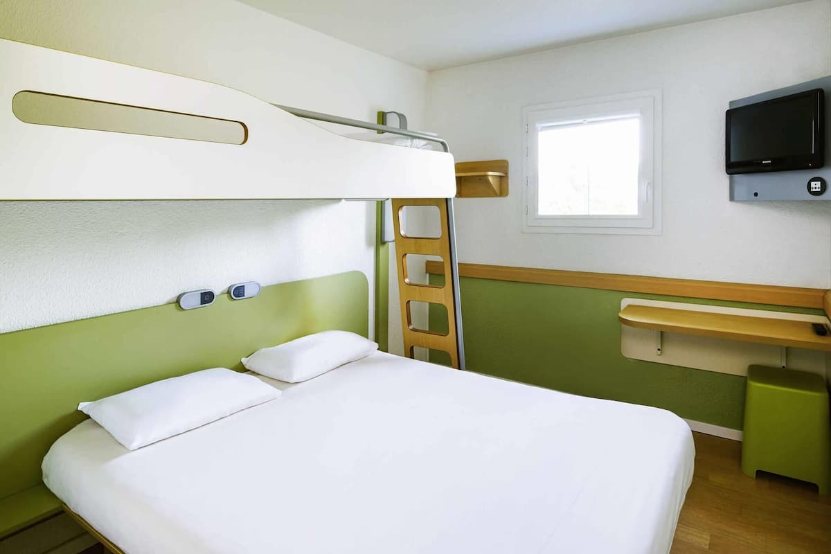 Una cama individual doble con un panel de control, un escritorio con silla, un televisor montado en la pared y una ventana.