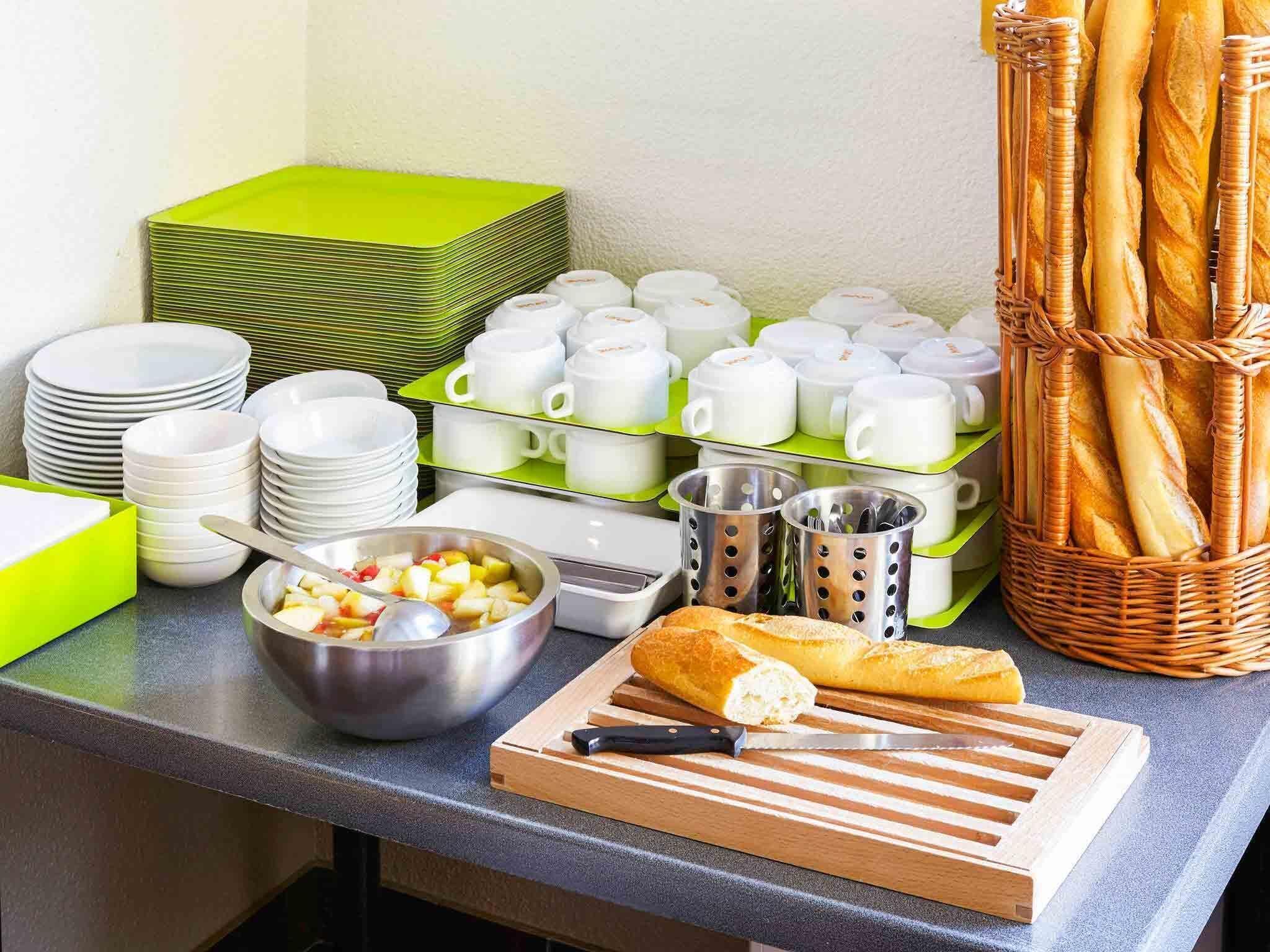 Disposição de buffet com pratos, copos, uma tigela de frutas, pão e talheres.