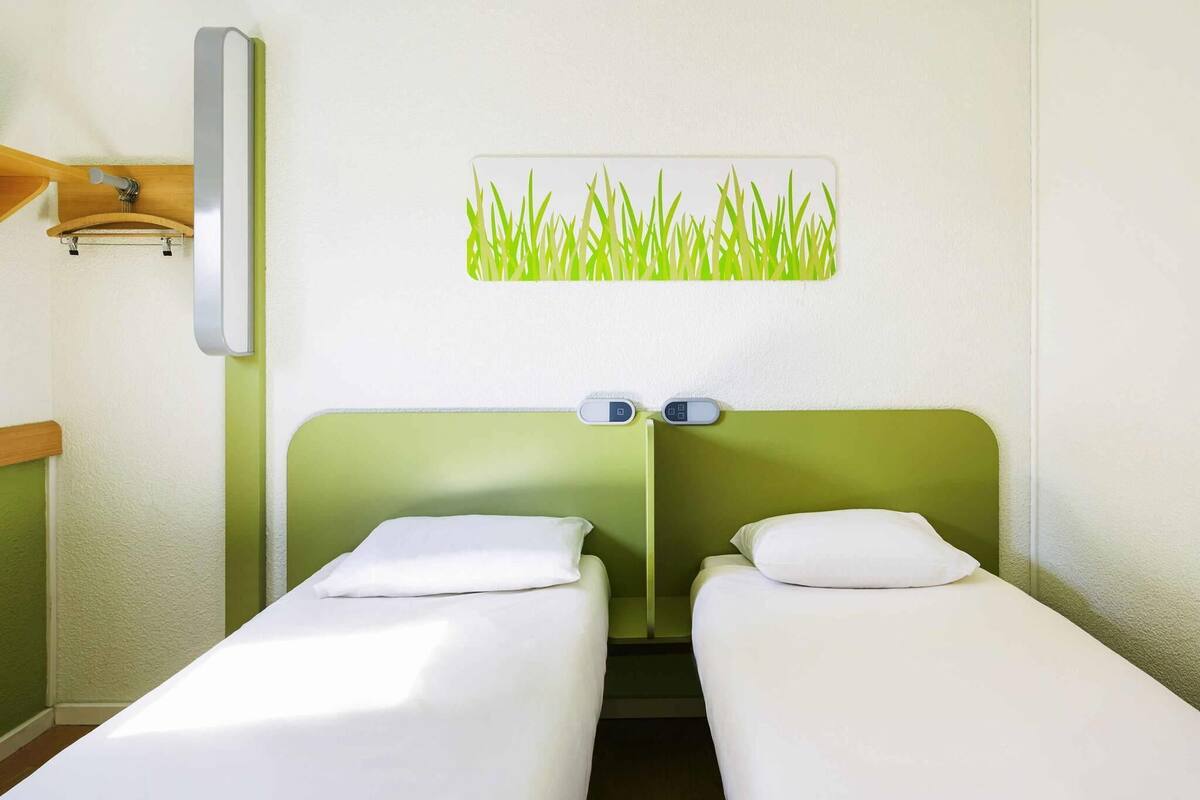 Habitación de hotel con dos camas individuales, cabecero verde, luz instalada en la pared y una obra de arte con motivos de hierba.