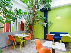 A modern indoor dining area with colorful chairs, white tables, and a large tree. - ibis budget Cavaillon (Cavaillon)